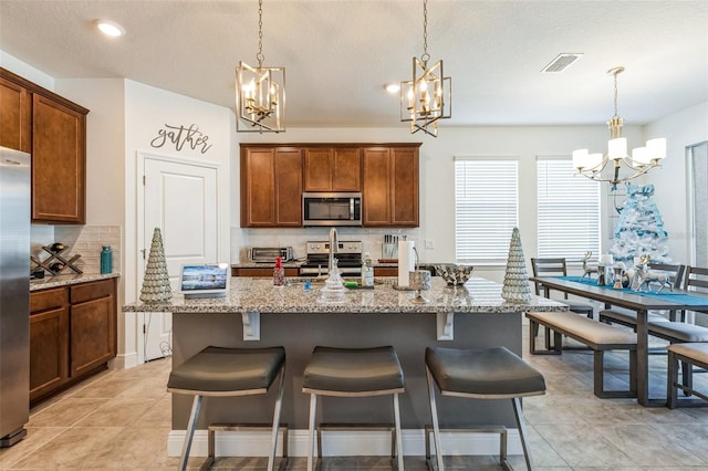 kitchen with a center island with sink, hanging light fixtures, light stone countertops, tasteful backsplash, and stainless steel appliances