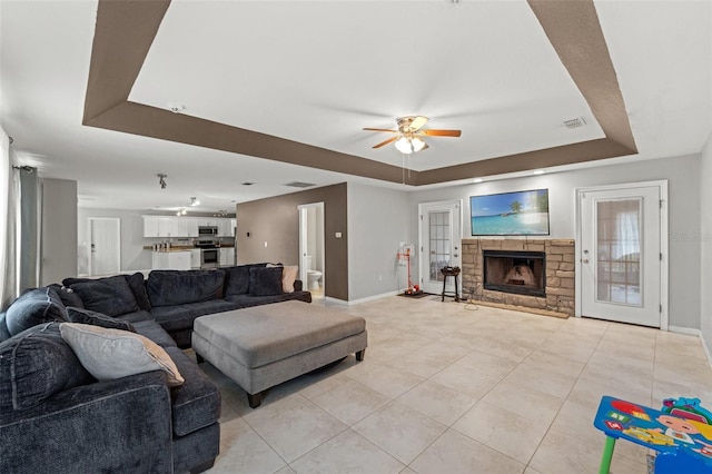 tiled living room with ceiling fan, a stone fireplace, and a raised ceiling