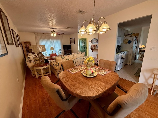 dining space with ceiling fan with notable chandelier, baseboards, a textured ceiling, light wood finished floors, and visible vents