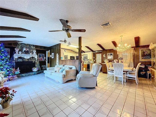 tiled living room featuring ceiling fan with notable chandelier, a textured ceiling, vaulted ceiling with beams, a stone fireplace, and wood walls