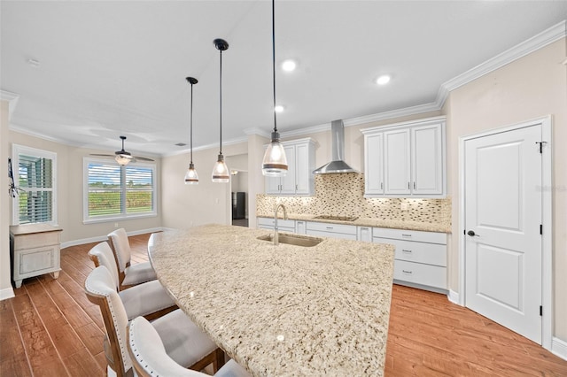 kitchen featuring sink, wall chimney exhaust hood, ceiling fan, an island with sink, and white cabinetry