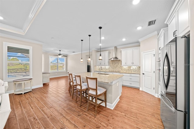 kitchen with stainless steel refrigerator, white cabinetry, wall chimney exhaust hood, pendant lighting, and a center island with sink