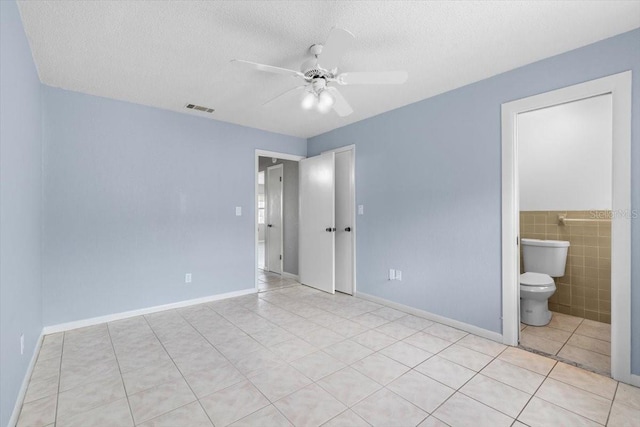 unfurnished bedroom featuring a textured ceiling, ceiling fan, light tile patterned floors, tile walls, and connected bathroom