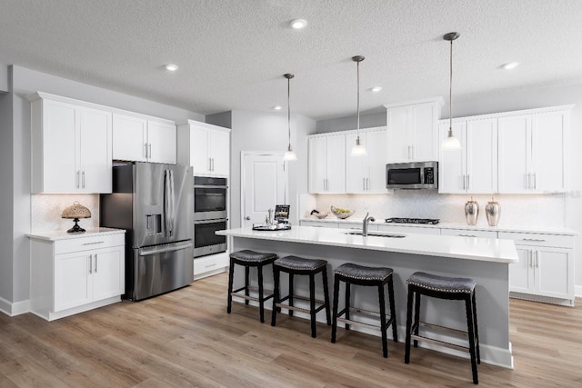 kitchen with hanging light fixtures, light hardwood / wood-style flooring, an island with sink, appliances with stainless steel finishes, and white cabinetry