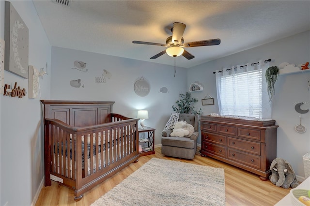 bedroom with a textured ceiling, light wood-type flooring, a nursery area, and ceiling fan