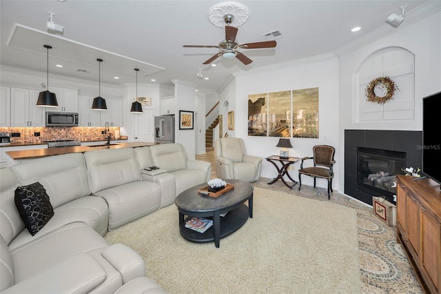 living room featuring a tiled fireplace, ceiling fan, crown molding, and sink