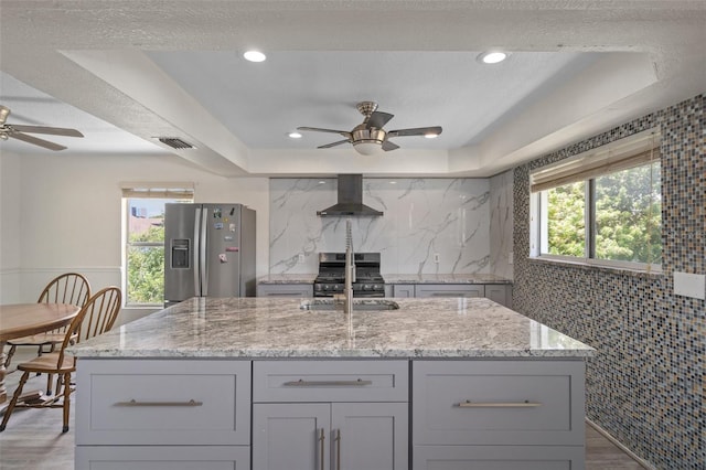 kitchen featuring appliances with stainless steel finishes, light stone counters, a raised ceiling, wall chimney range hood, and a kitchen island