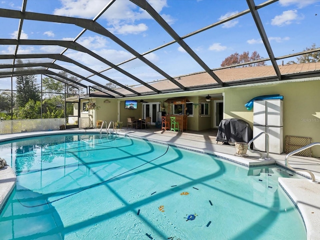 view of swimming pool featuring exterior bar, ceiling fan, glass enclosure, a patio, and a grill