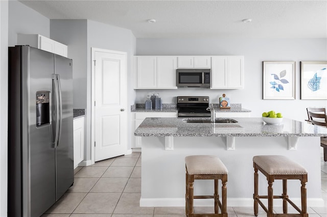 kitchen with light stone counters, stainless steel appliances, light tile patterned floors, white cabinets, and an island with sink