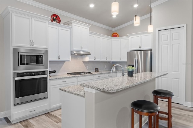 kitchen with a kitchen island with sink, ornamental molding, appliances with stainless steel finishes, light stone counters, and white cabinetry