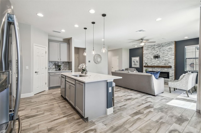 kitchen with stainless steel appliances, sink, a center island with sink, gray cabinets, and a stone fireplace