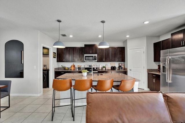 kitchen with dark brown cabinetry, pendant lighting, a kitchen island with sink, light tile patterned flooring, and appliances with stainless steel finishes