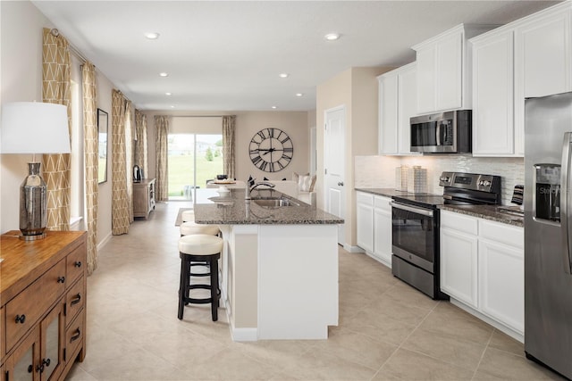 kitchen featuring backsplash, dark stone countertops, an island with sink, white cabinetry, and stainless steel appliances