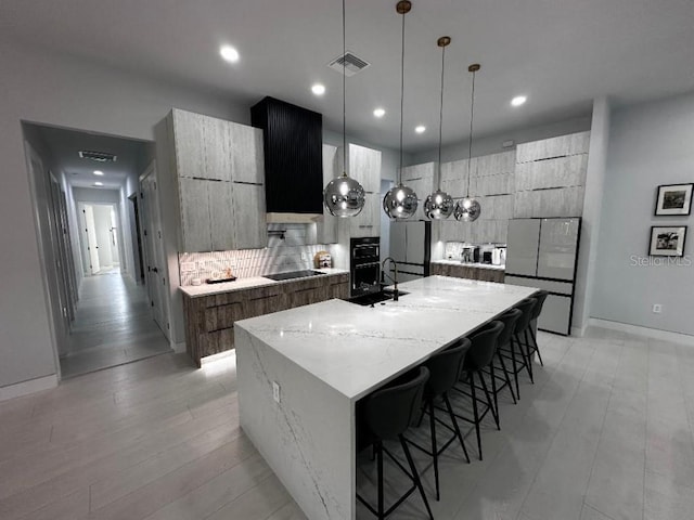 kitchen featuring hanging light fixtures, light stone counters, backsplash, a large island with sink, and a kitchen bar