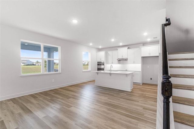 kitchen with a kitchen bar, stainless steel double oven, light hardwood / wood-style floors, white cabinetry, and an island with sink