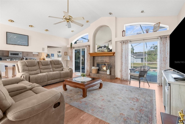 living room with high vaulted ceiling, a tiled fireplace, ceiling fan, and light hardwood / wood-style flooring