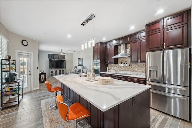 kitchen featuring wall chimney exhaust hood, decorative light fixtures, an island with sink, stainless steel fridge, and a breakfast bar area