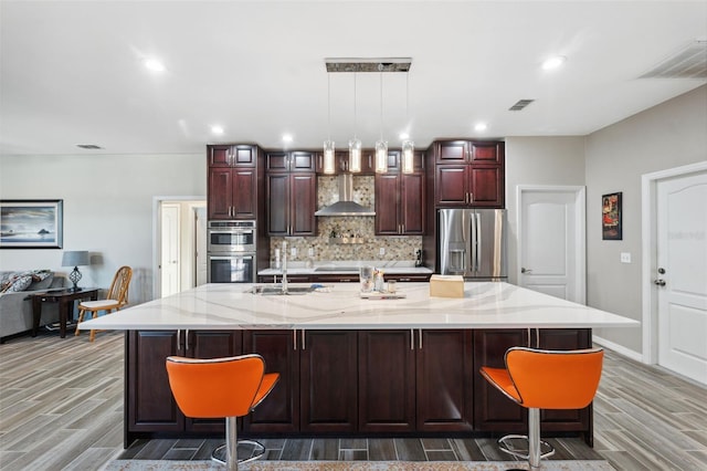 kitchen featuring stainless steel appliances, hardwood / wood-style flooring, a large island with sink, hanging light fixtures, and wall chimney range hood