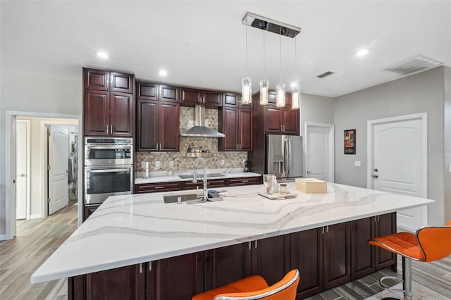 kitchen with stainless steel appliances, decorative light fixtures, wall chimney exhaust hood, backsplash, and a kitchen island with sink