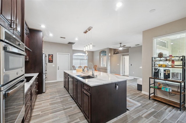 kitchen featuring decorative light fixtures, stainless steel appliances, a kitchen island with sink, ceiling fan, and sink