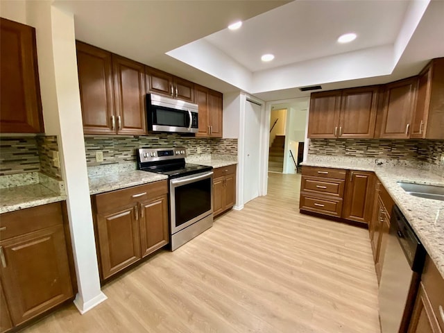 kitchen with light stone counters, stainless steel appliances, tasteful backsplash, light wood-type flooring, and a tray ceiling