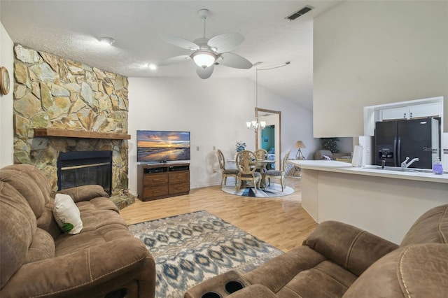 living room with ceiling fan with notable chandelier, a textured ceiling, a stone fireplace, vaulted ceiling, and light hardwood / wood-style flooring