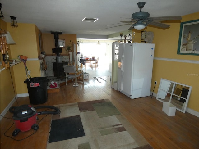 kitchen with a ceiling fan, white refrigerator with ice dispenser, wood finished floors, baseboards, and a wood stove