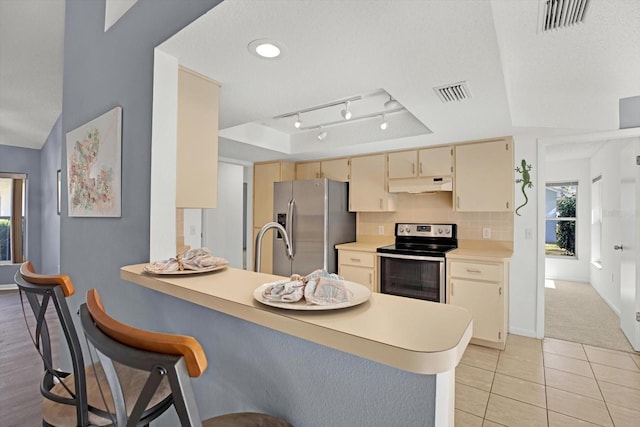 kitchen with kitchen peninsula, decorative backsplash, a tray ceiling, stainless steel appliances, and cream cabinetry