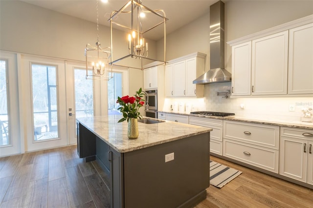 kitchen with white cabinets, wall chimney exhaust hood, decorative light fixtures, stainless steel appliances, and a kitchen island with sink