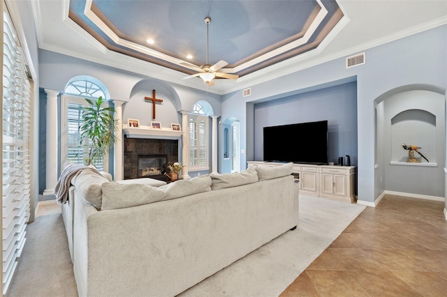 tiled living room featuring ornate columns, crown molding, a fireplace, ceiling fan, and a tray ceiling