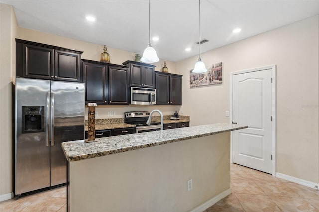 kitchen with light tile patterned floors, appliances with stainless steel finishes, pendant lighting, light stone countertops, and a kitchen island with sink