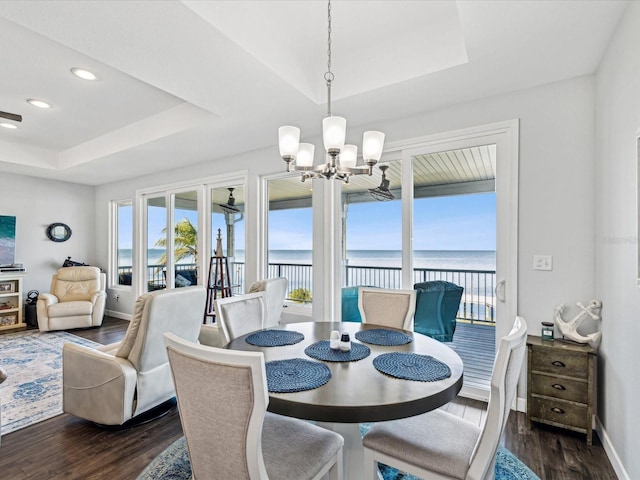 dining space with a notable chandelier, a tray ceiling, dark wood-type flooring, and a water view