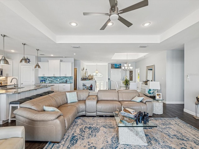 living room with sink, ceiling fan with notable chandelier, dark wood-type flooring, and a raised ceiling