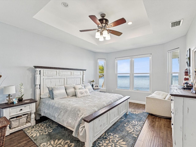 bedroom featuring a water view, a tray ceiling, dark hardwood / wood-style flooring, ceiling fan, and multiple windows