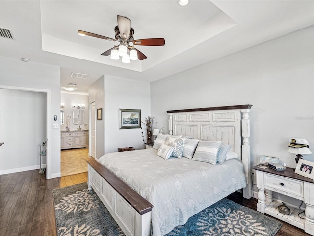 bedroom with hardwood / wood-style floors, a tray ceiling, ensuite bath, and ceiling fan