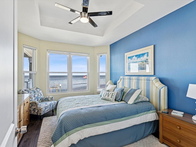 bedroom featuring a raised ceiling, wood-type flooring, a water view, and ceiling fan