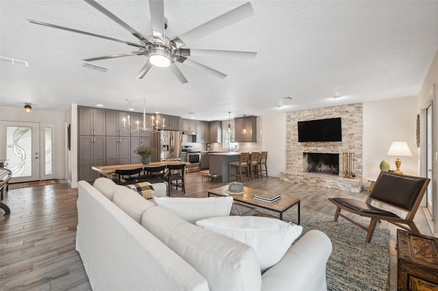 living room with a stone fireplace, ceiling fan with notable chandelier, a textured ceiling, and light hardwood / wood-style flooring