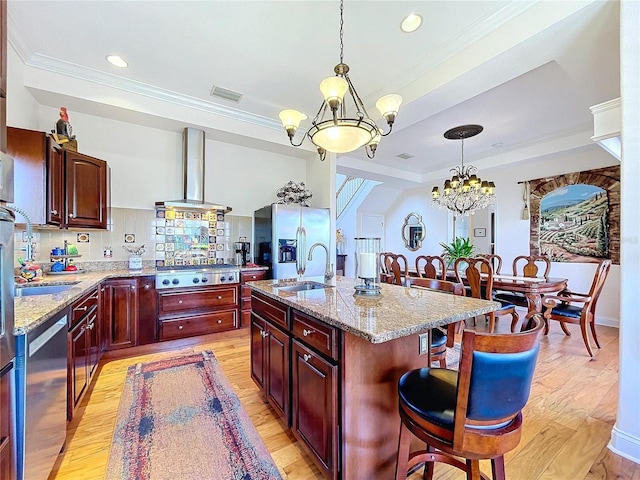 kitchen with a kitchen bar, an island with sink, a notable chandelier, stainless steel appliances, and wall chimney range hood