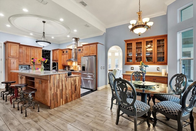 kitchen featuring light stone counters, hanging light fixtures, a center island with sink, stainless steel fridge, and range hood