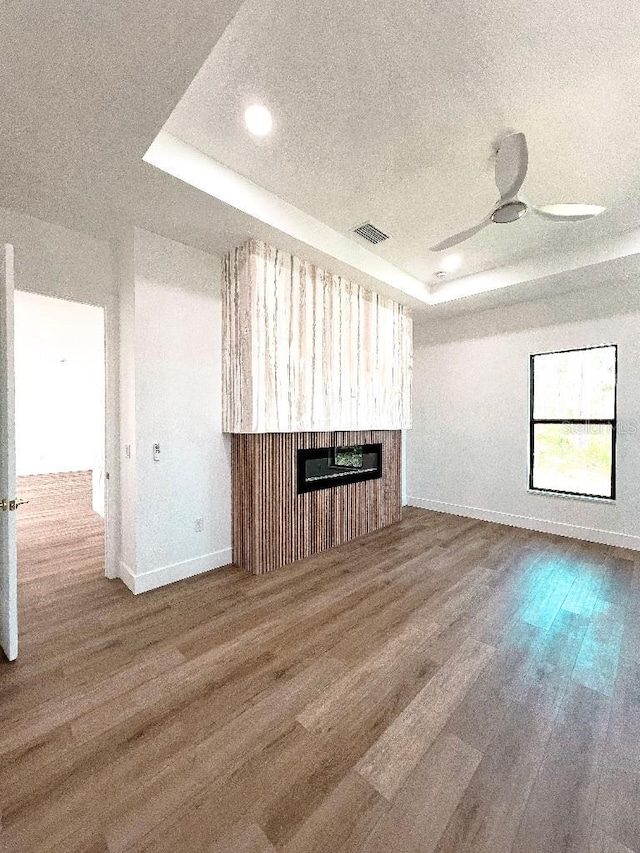 unfurnished living room with ceiling fan, a tray ceiling, wood finished floors, and a glass covered fireplace