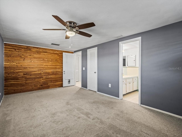 unfurnished bedroom with ceiling fan, light colored carpet, ensuite bathroom, and wooden walls