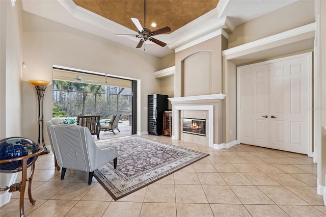 tiled living room featuring a tile fireplace, ceiling fan, a high ceiling, ornamental molding, and a raised ceiling