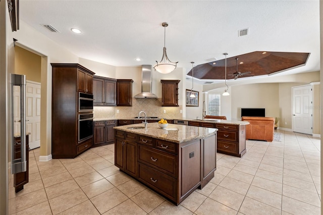 kitchen with decorative light fixtures, an island with sink, sink, a tray ceiling, and wall chimney exhaust hood