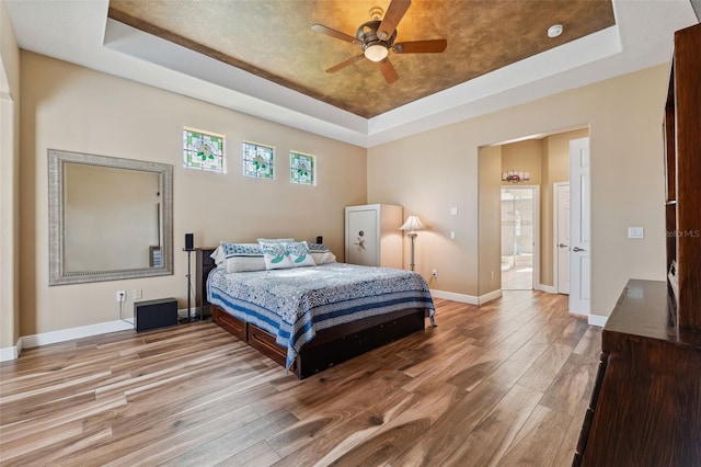 bedroom with a raised ceiling, ceiling fan, connected bathroom, and light hardwood / wood-style floors