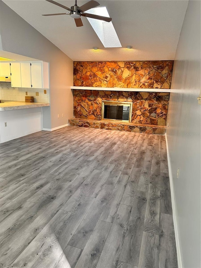 unfurnished living room featuring ceiling fan, a stone fireplace, vaulted ceiling with skylight, and light wood-type flooring