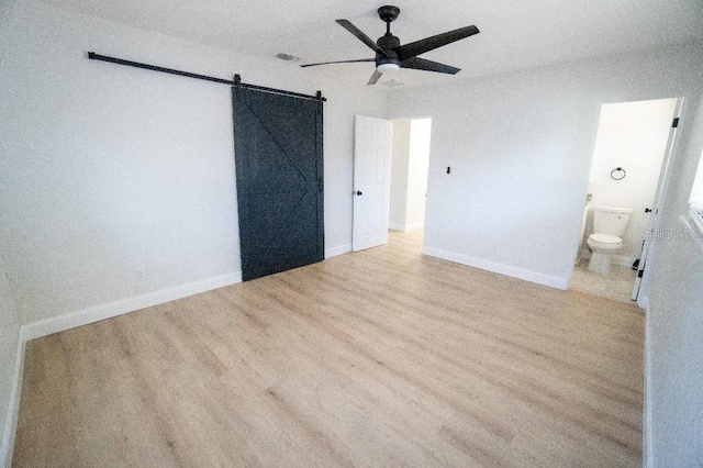 unfurnished bedroom featuring ceiling fan, a barn door, light hardwood / wood-style floors, and ensuite bath