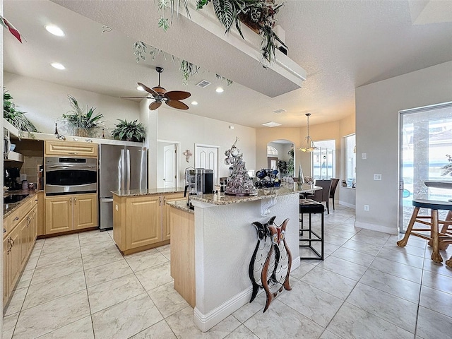 kitchen with light brown cabinetry, hanging light fixtures, an island with sink, stainless steel appliances, and light stone countertops