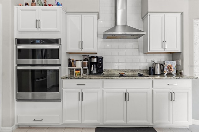 kitchen with double oven, white cabinets, backsplash, light stone countertops, and wall chimney range hood