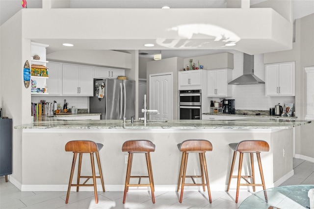 kitchen with wall chimney range hood, light stone countertops, white cabinets, and appliances with stainless steel finishes