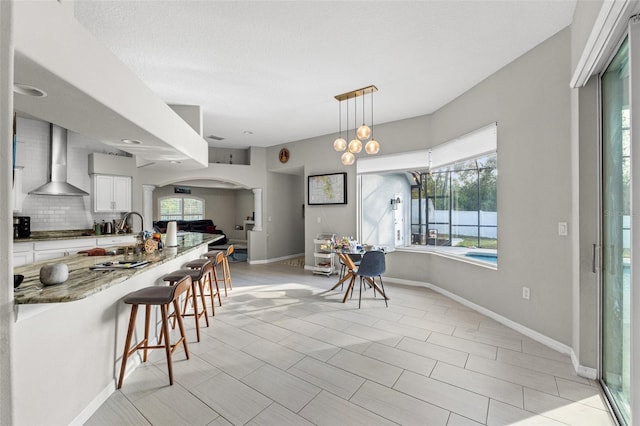 kitchen featuring wall chimney range hood, a breakfast bar, white cabinetry, hanging light fixtures, and light stone counters